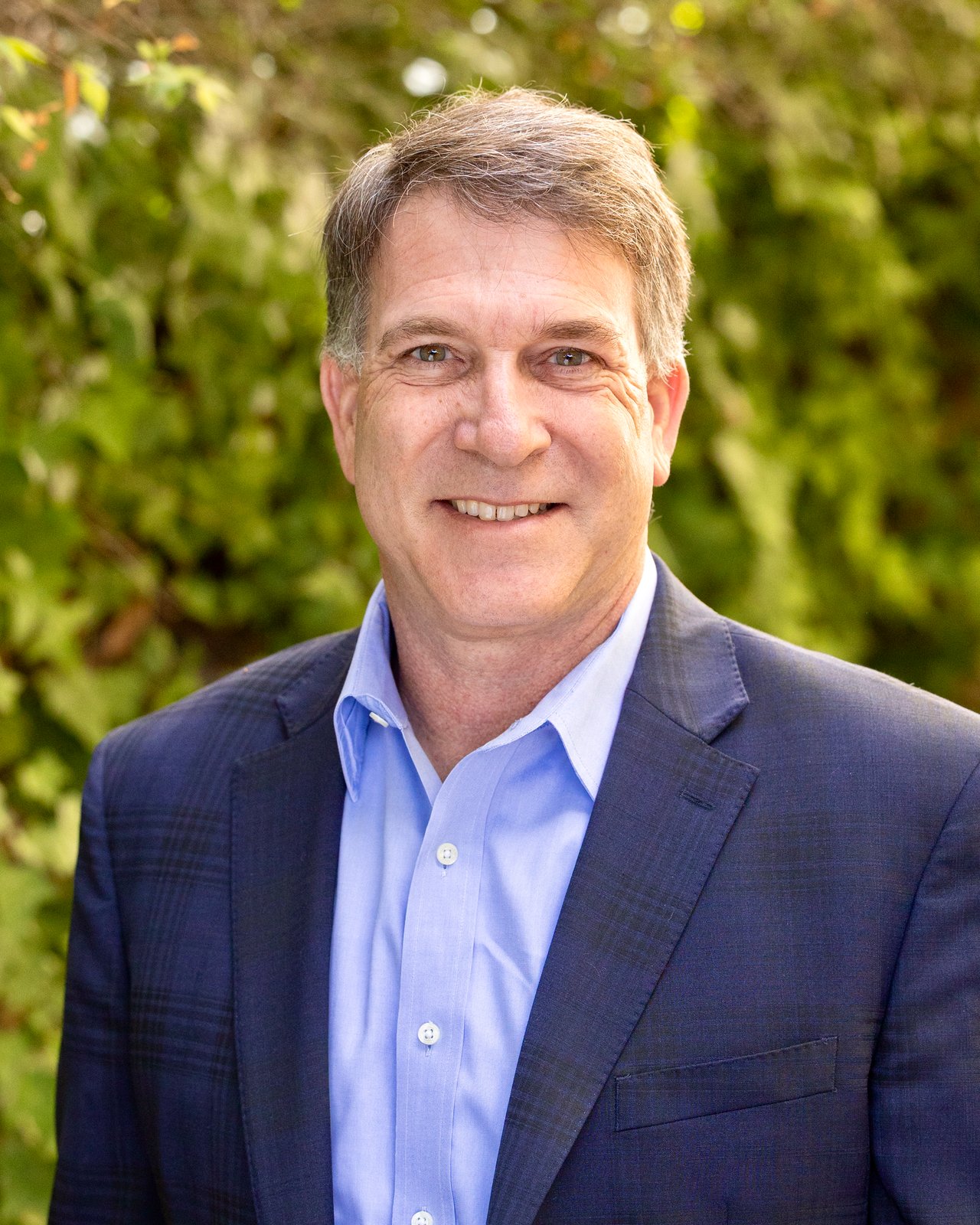 Adam Hamalian smiling in a navy suit jacket against leafy greenery.