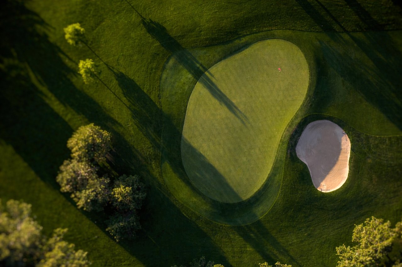 Aerial golf green and sand bunker crossed by long shadows in warm evening light.