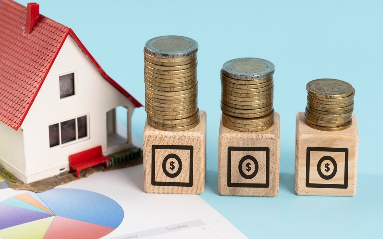 Miniature house beside three coin stacks on dollar-marked blocks and a partially visible pie chart.