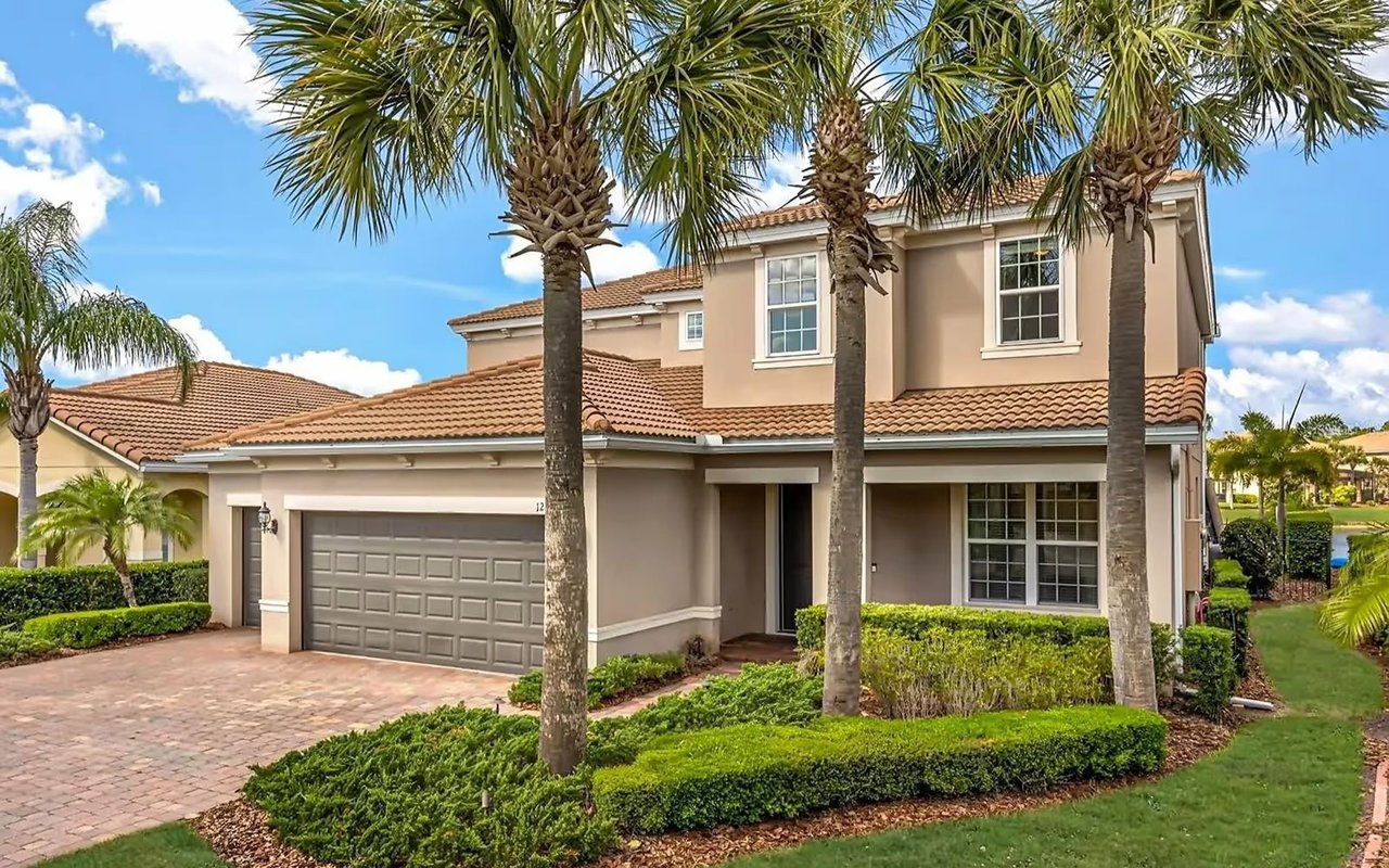 Two-story beige home with a tile roof, double garage, and tall palms in the front yard.