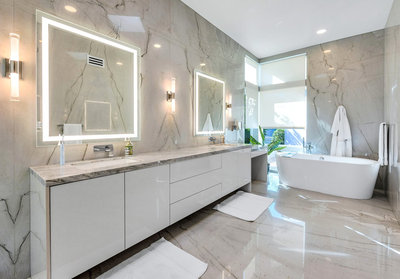 Modern marble bathroom with a long double vanity, illuminated mirrors, and a freestanding tub.