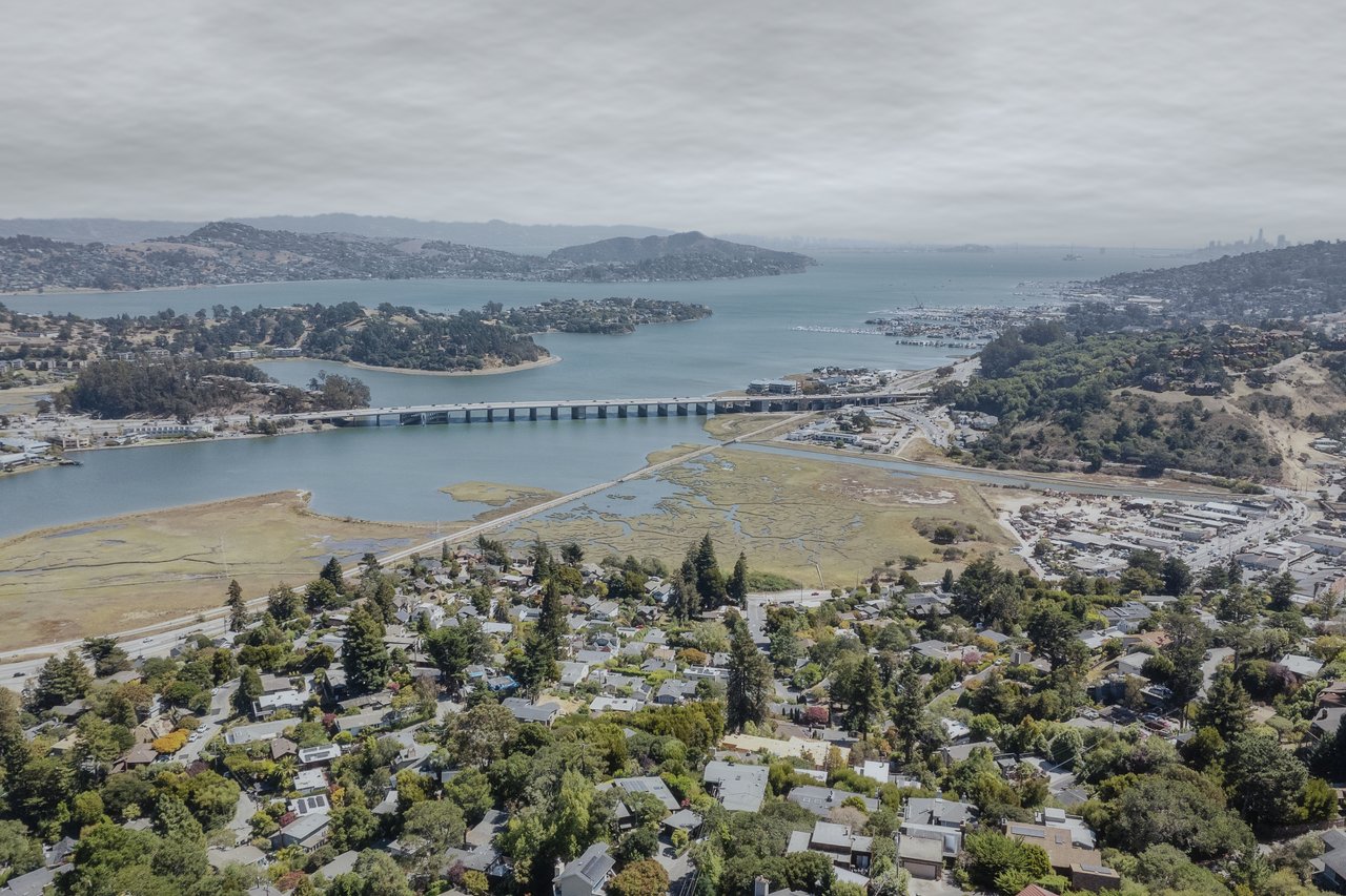 Aerial view of a Bay Area inlet, bridge, marshland, and hillside neighborhoods beneath cloudy skies.