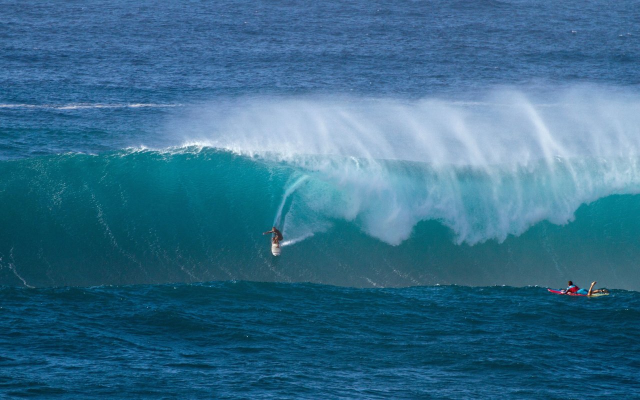 Surfer descends a towering turquoise wave as two people watch from the water.