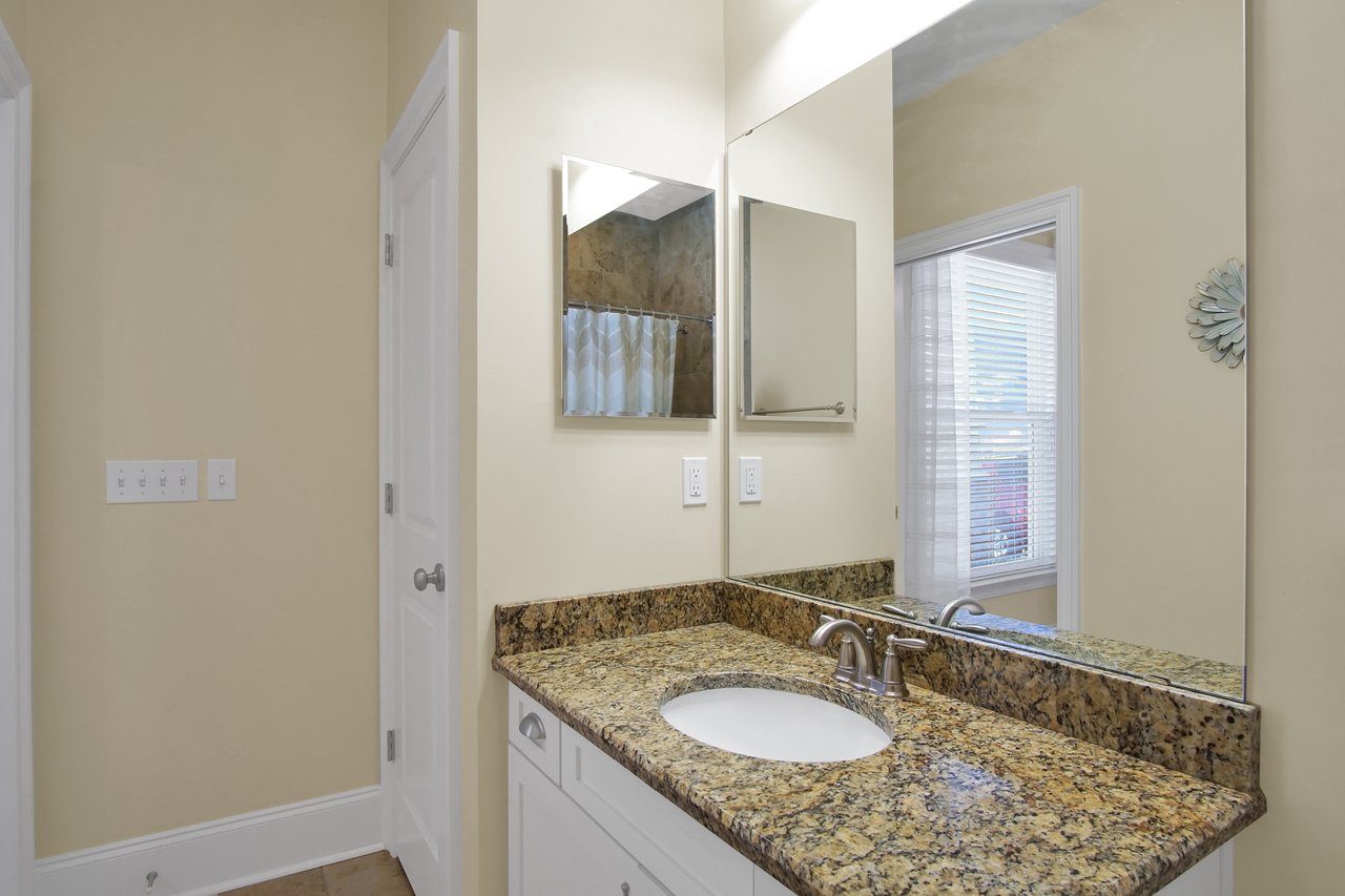 Clean bathroom with beige walls, a granite countertop, and a round sink. A large mirror reflects natural light from a window with blinds, creating a bright atmosphere.