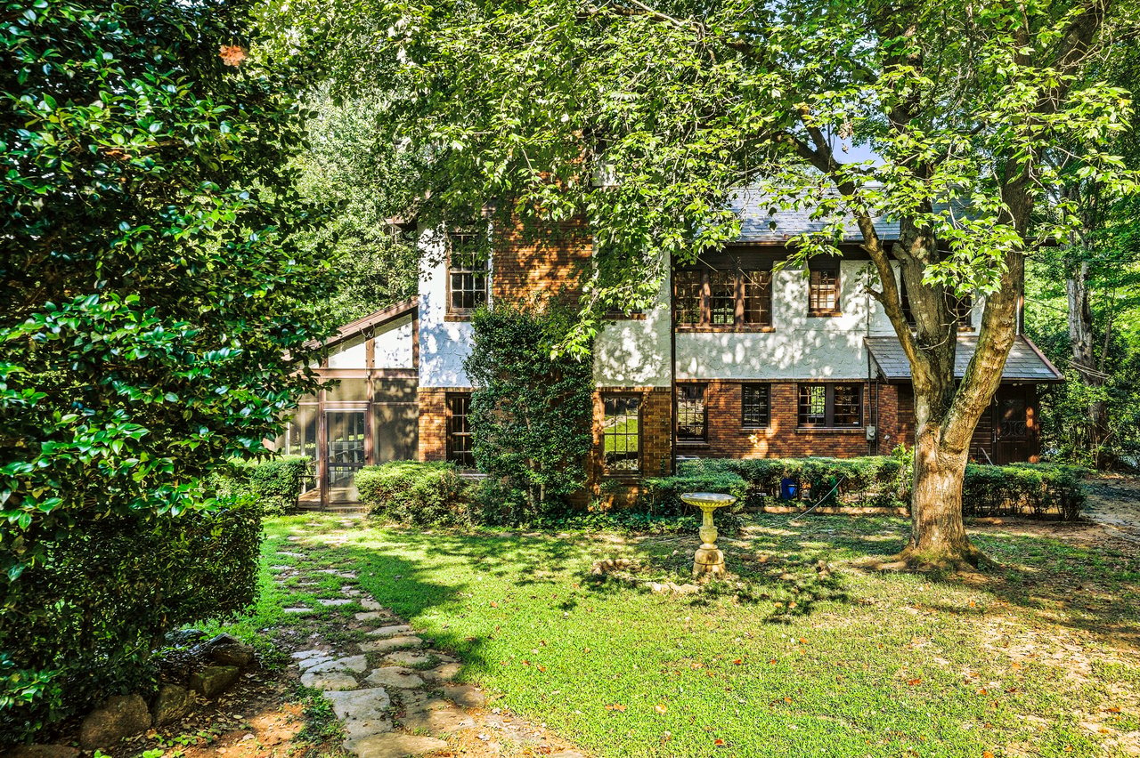 Two-story brick and stucco house framed by mature trees, with a stone garden path and birdbath.