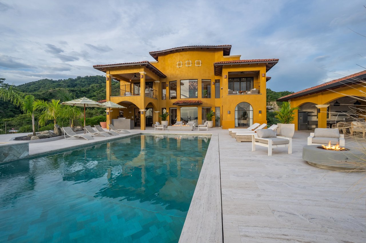 Ochre villa behind a turquoise pool, with a furnished terrace and forested hills beyond.