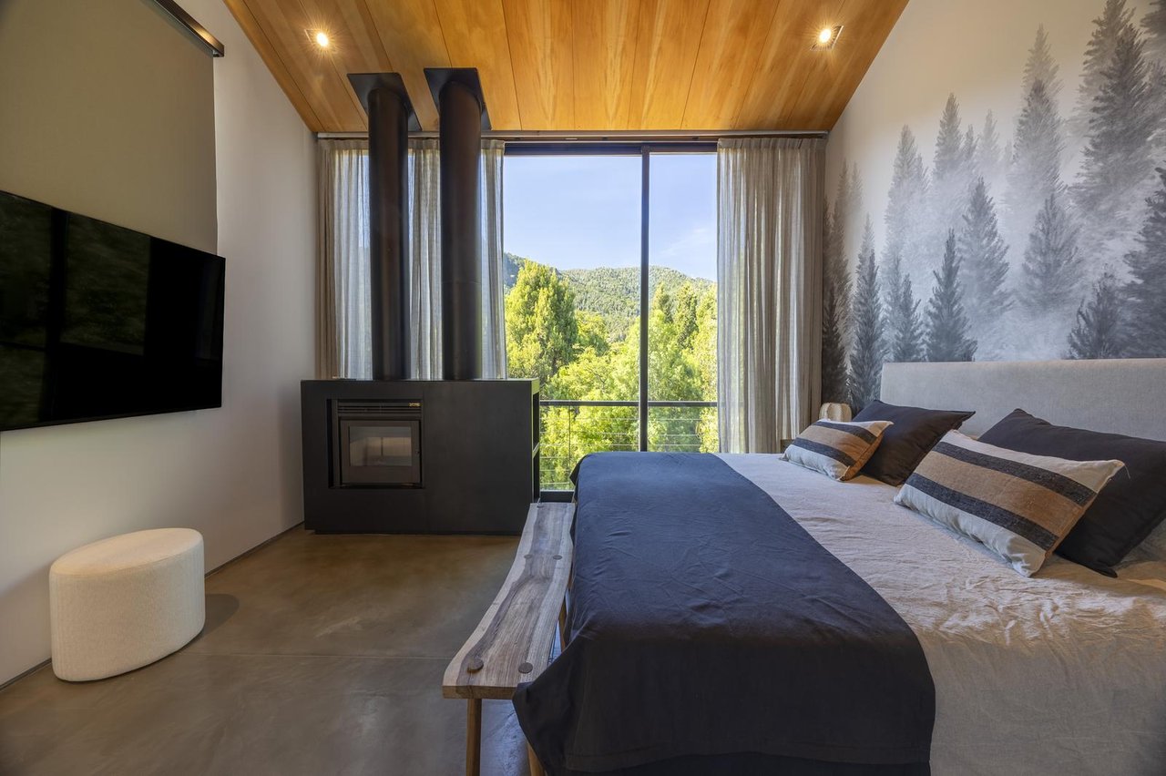 Bedroom with a modern fireplace and large window overlooking green trees and a mountain.