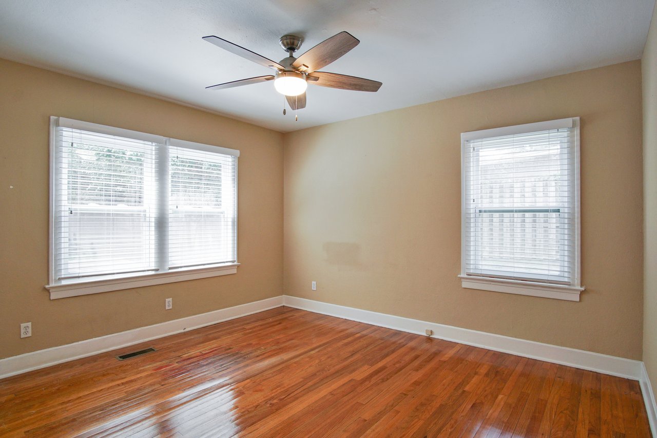 Empty room with wooden floors, beige walls, and two large windows with blinds. A ceiling fan with a light is centered on the ceiling. Bright and airy atmosphere.