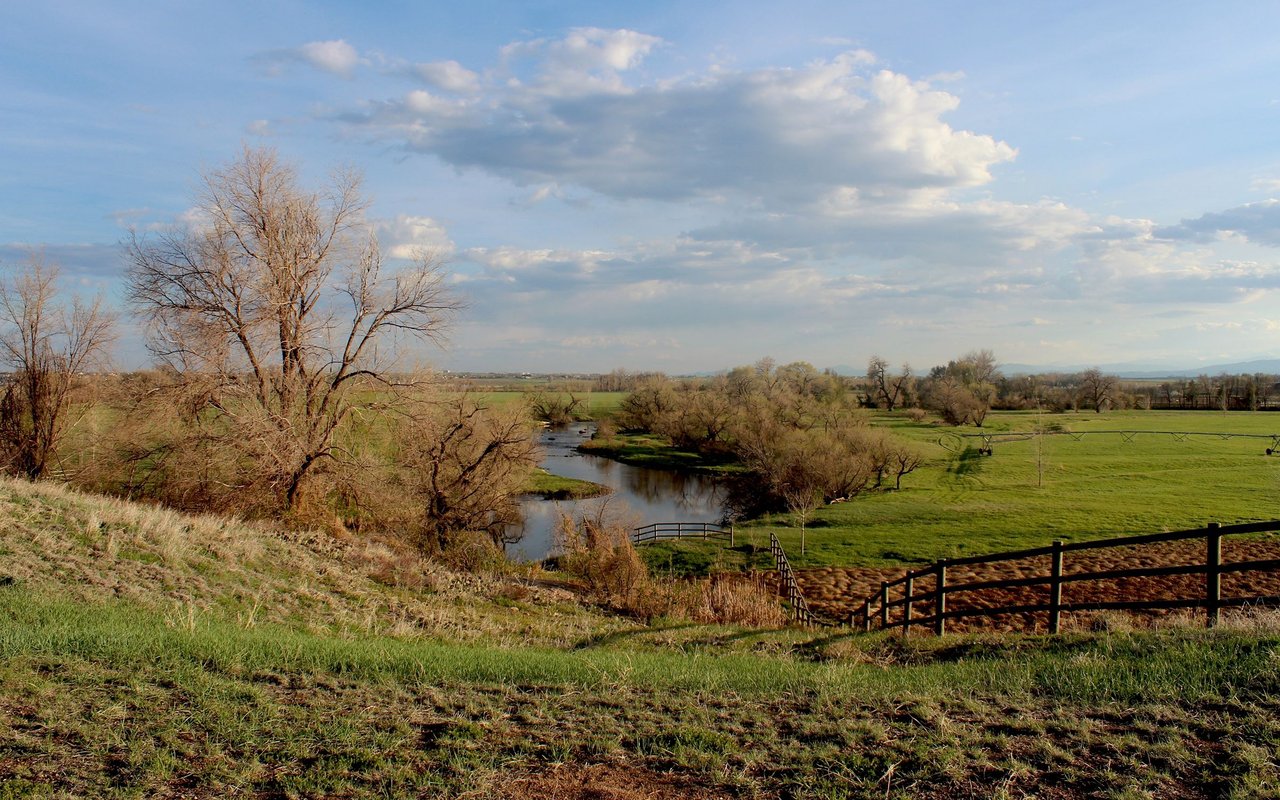 Rural creek winding through green fields, with leafless trees and a black wooden fence.