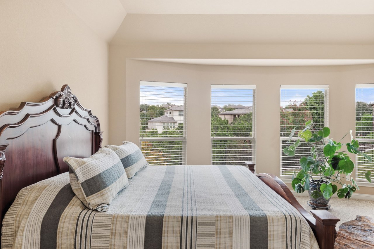 Bedroom with an ornate wood headboard, striped bedding, and windows overlooking trees and neighboring homes.