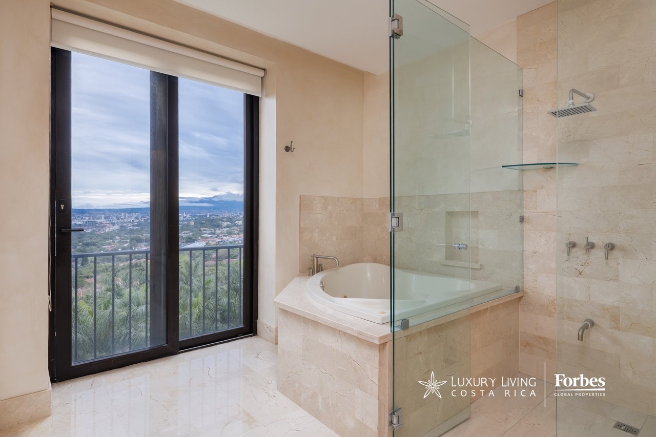 Marble penthouse bathroom with a glass shower, soaking tub, and balcony overlooking trees and the city.