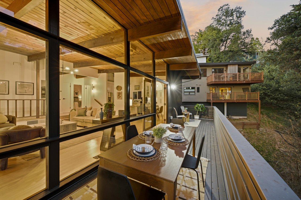 Dining deck beside a glass-walled living room, beneath a broad wood-plank overhang.