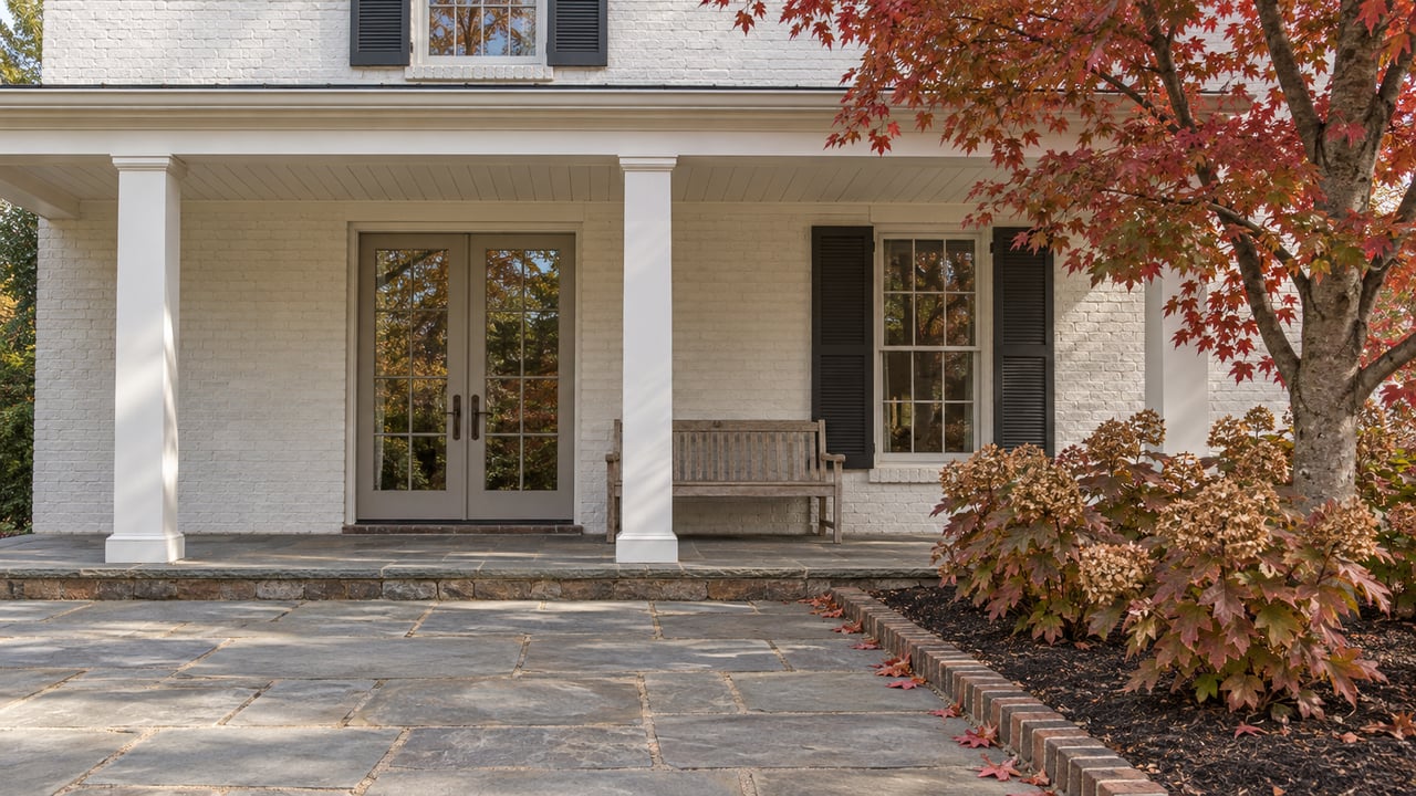 White painted-brick home with a sheltered porch, bluestone terrace, weathered teak bench and red autumn maple.
