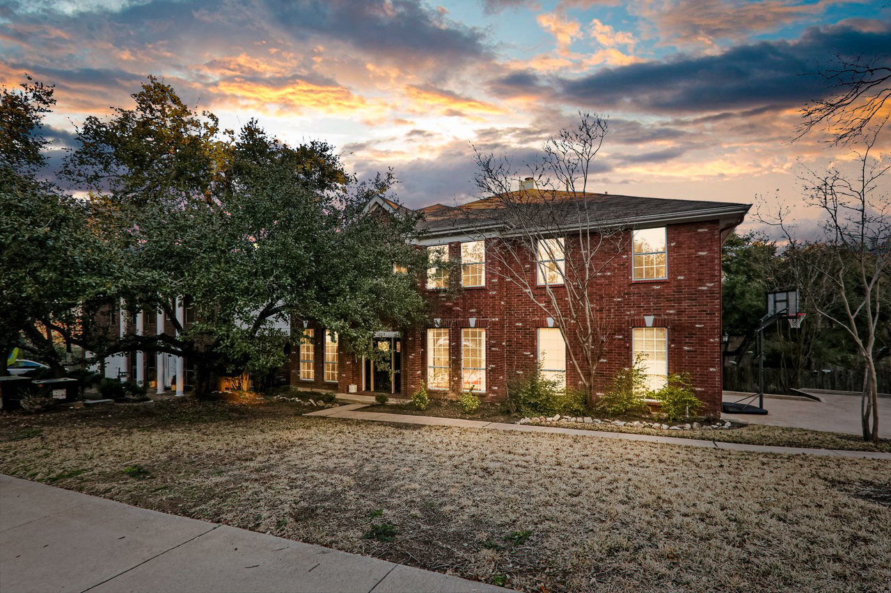 Two-story red-brick home with glowing windows and a basketball hoop beside the driveway at dusk.