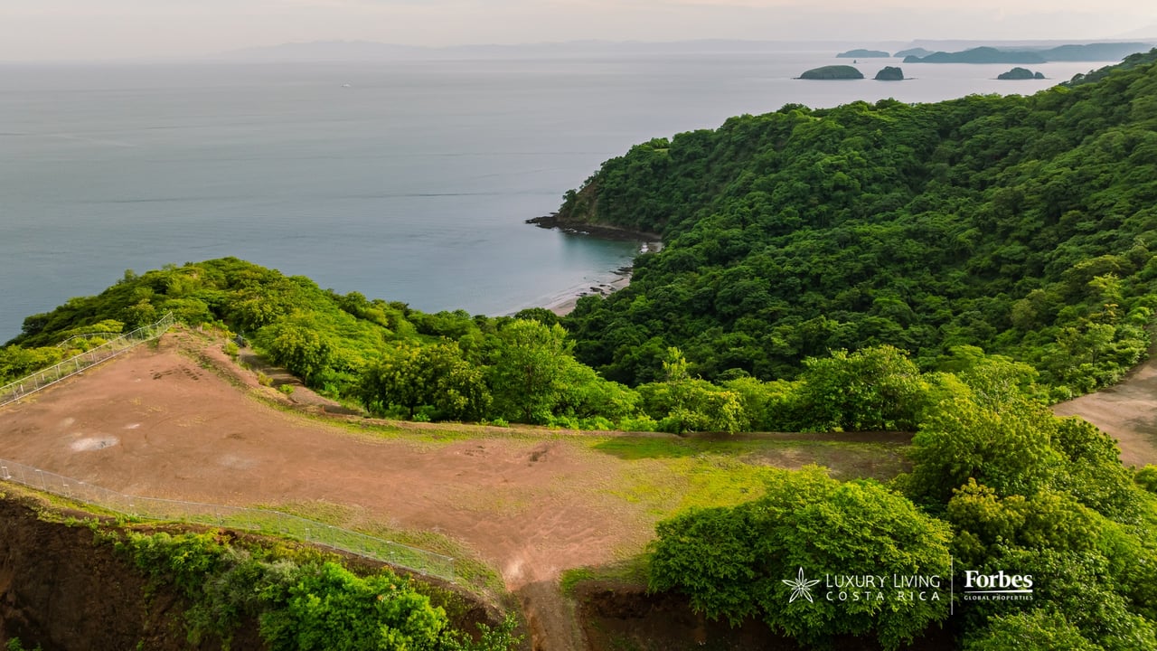 Aerial view of a cleared residential lot above a forested coast, with ocean and islands.