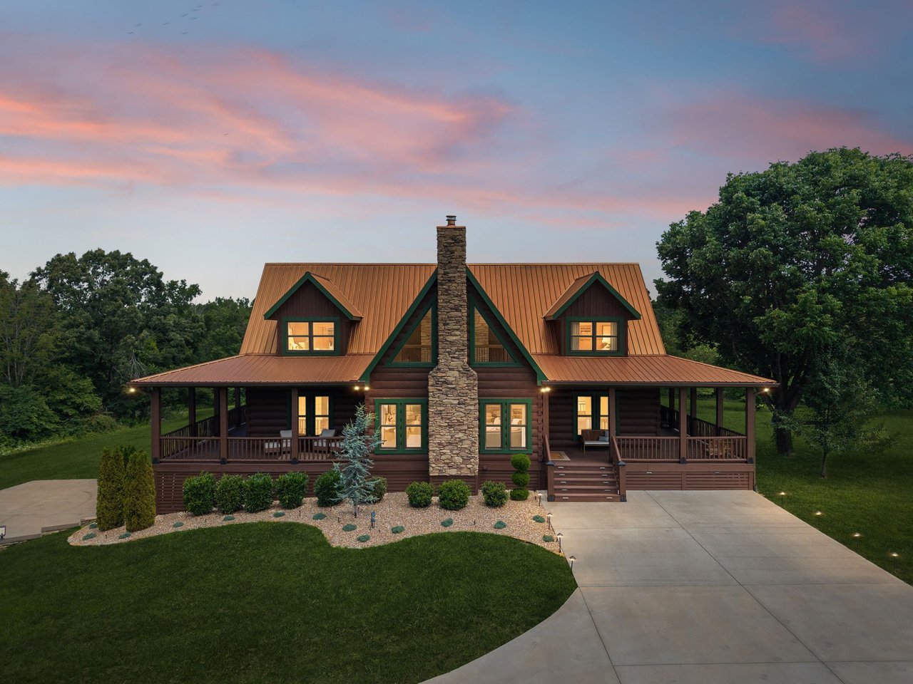 Log cabin with a stone chimney, copper-colored roof, and covered wraparound porch at dusk.