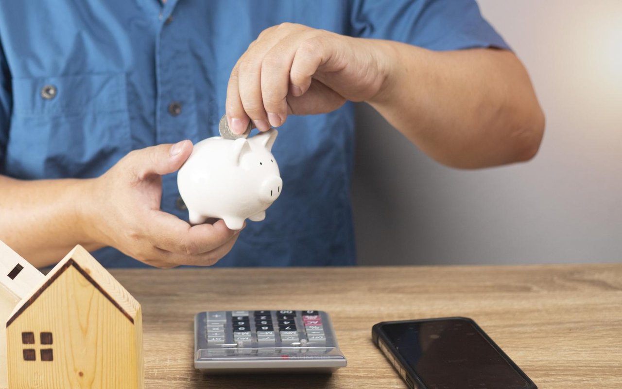 Person putting a coin into a piggy bank beside a wooden house model and calculator.