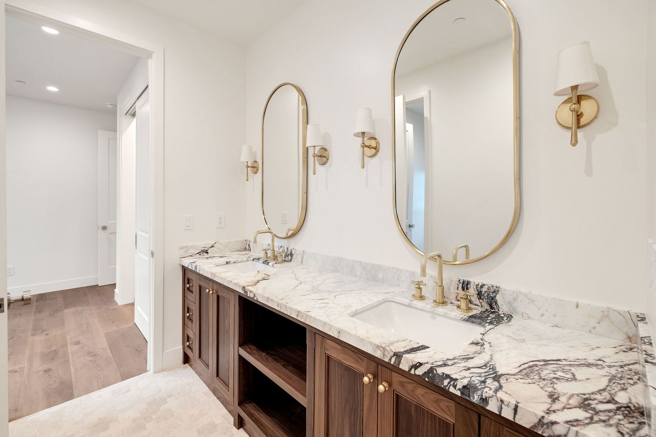Bathroom with a double marble vanity, dark wood cabinets, brass fixtures, and two arched mirrors.