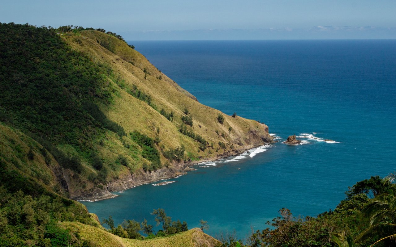 Green coastal hills meet a rocky shoreline beside the blue ocean.