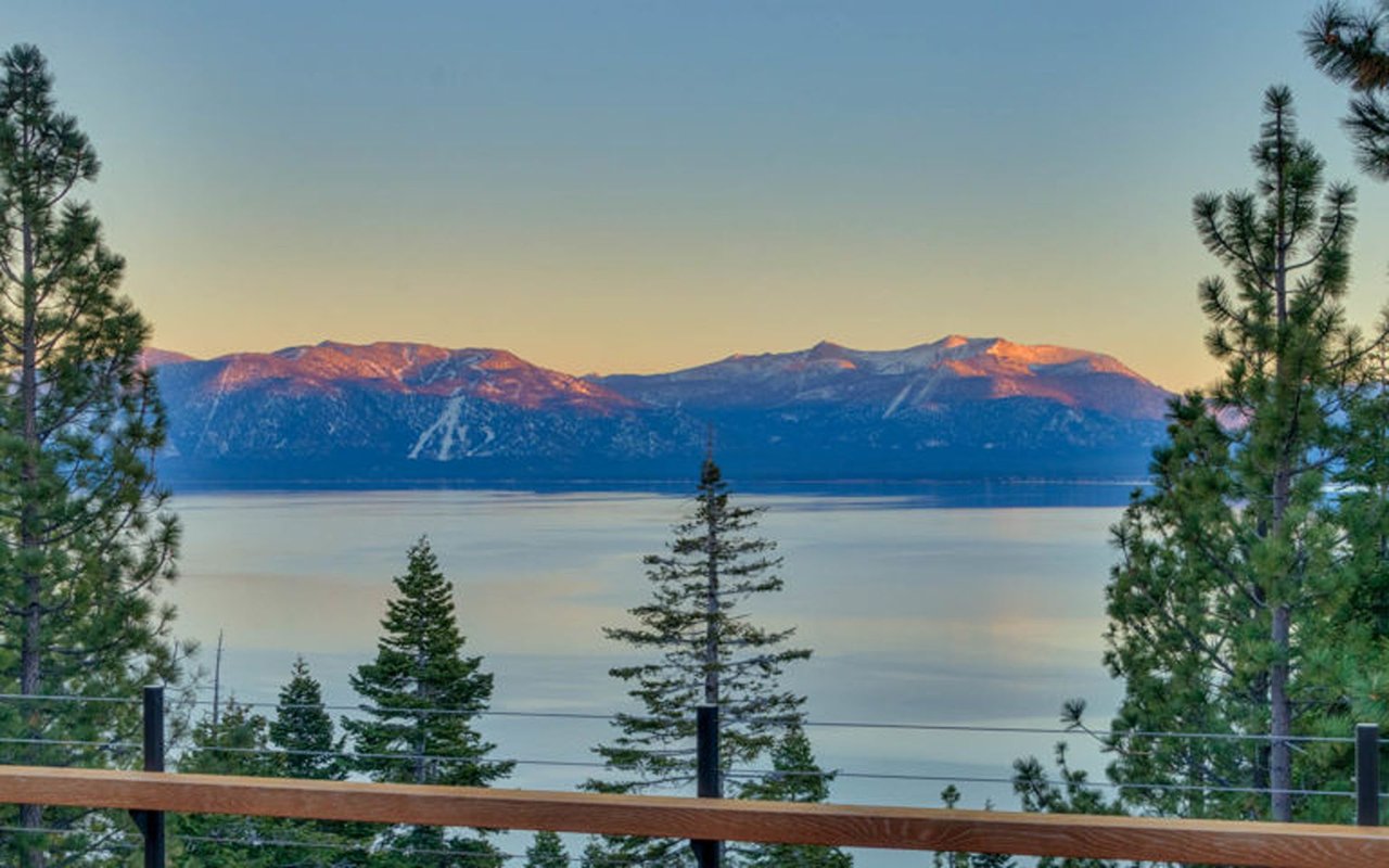 Lake Tahoe and distant mountains at golden hour, framed by pine trees above a deck railing.