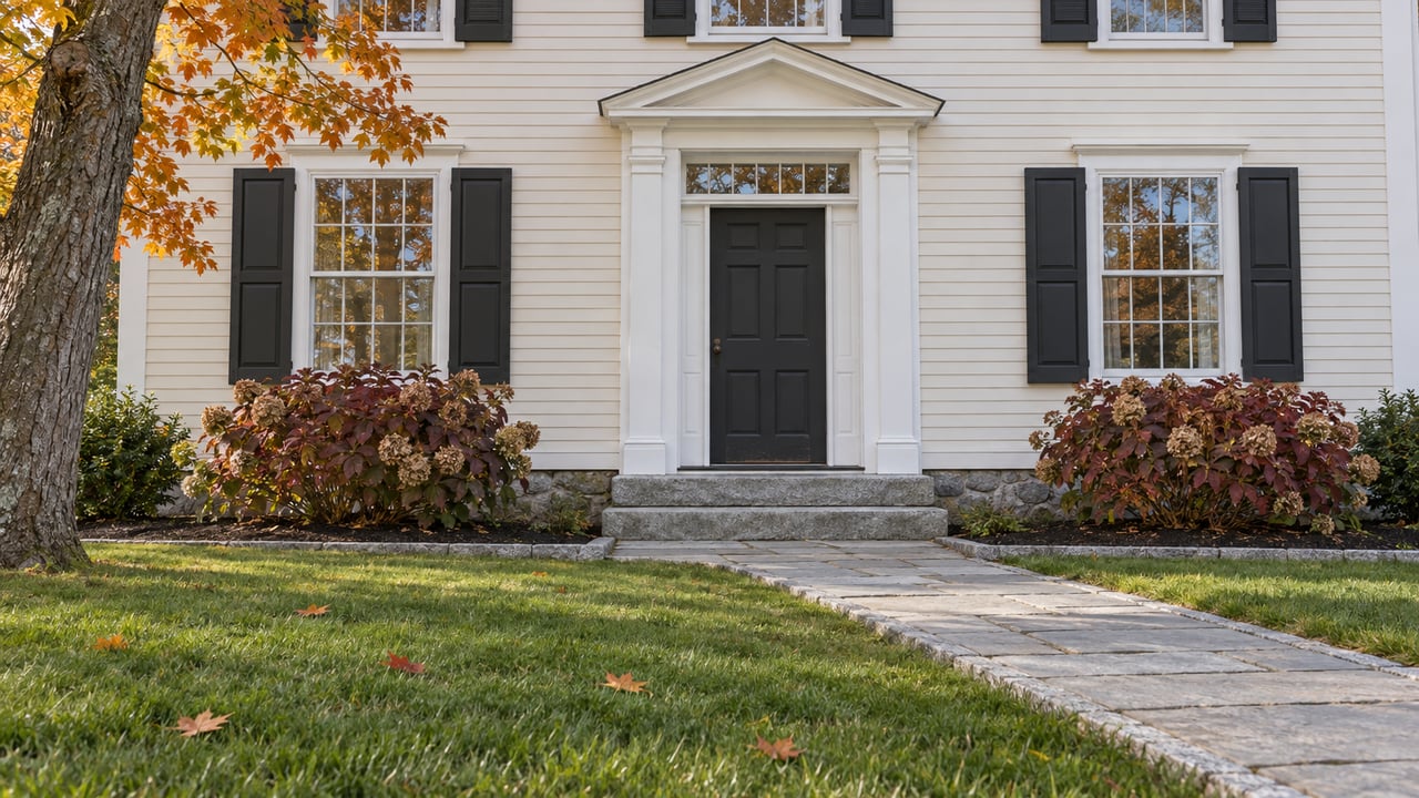 Autumn light on an ivory clapboard home with black shutters, a charcoal front door, granite steps and an amber-leaved maple beside the lawn.