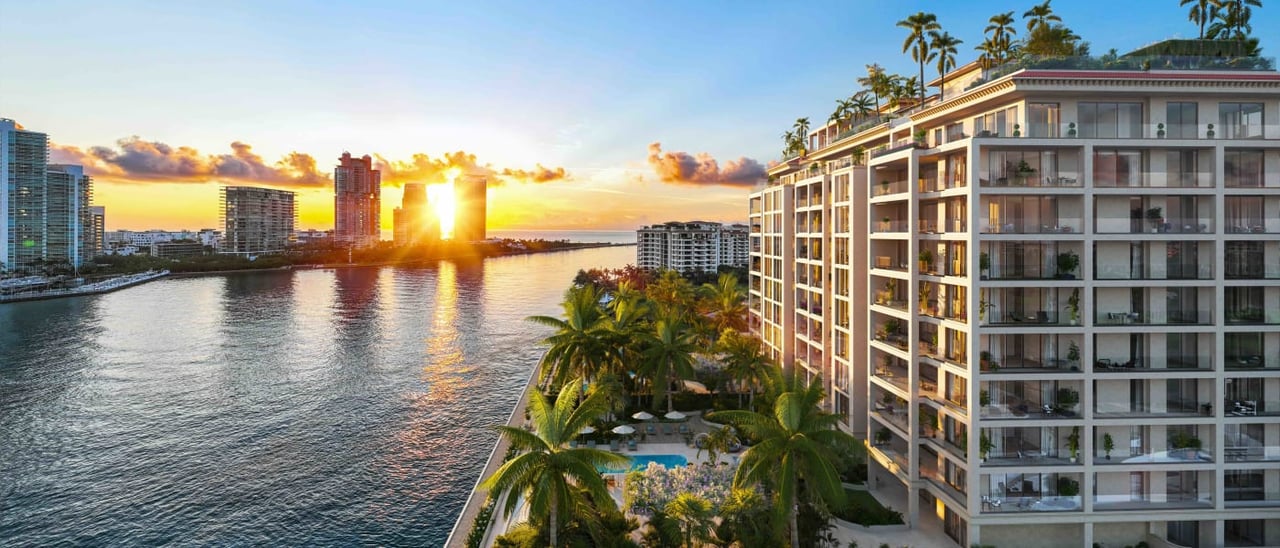 Waterfront condominium with glass balconies and palm-lined pool grounds overlooking the bay at sunset.