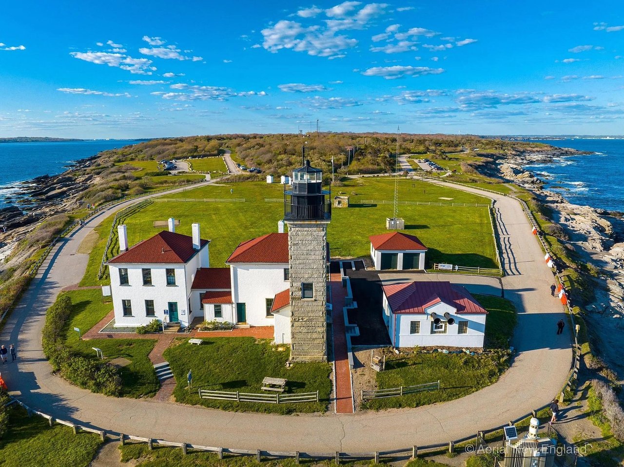 Aerial view of a coastal lighthouse complex surrounded by ocean, rocky shore, and looping paved paths.