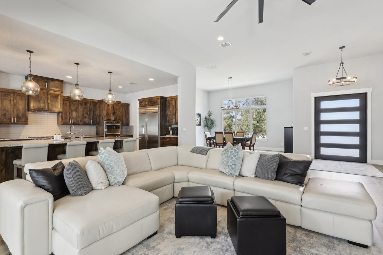 Large European sectional, white leather sofa in the forefront with a view of a chef's designed kitchen with custom hardwood stained cabinets with three pendant lights and stainlesss steel professional appliances. In addition in the background is a dining room with six seating table, three tall windows with hill country views and a view of a large custom metal pivot front door with five glass panels.