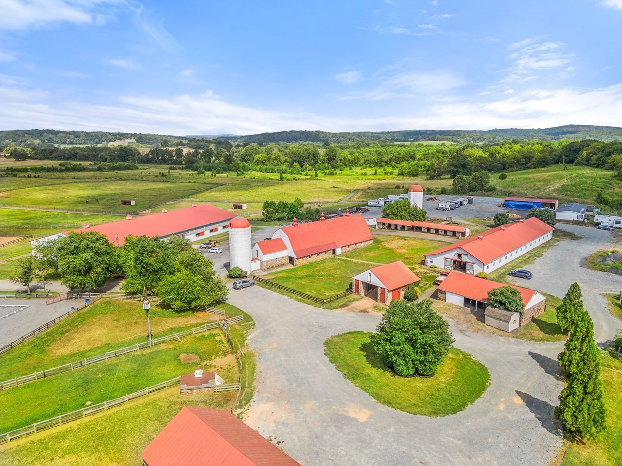 Aerial view of an equestrian property with red-roofed barns, fenced paddocks, and rolling countryside.