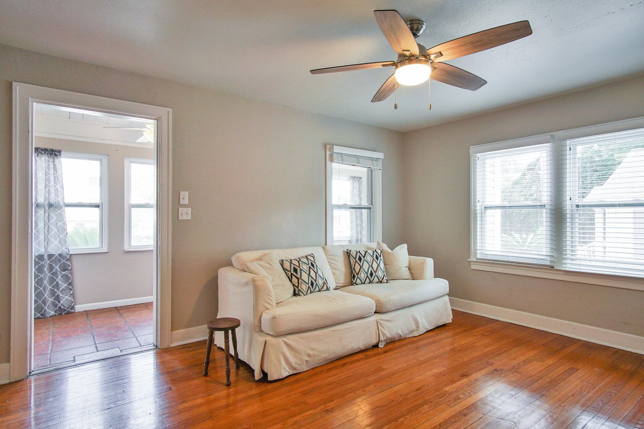 Bright living room with a white sofa and patterned cushions on a hardwood floor. Ceiling fan above, large windows with blinds, small stool nearby. Cozy and inviting atmosphere.