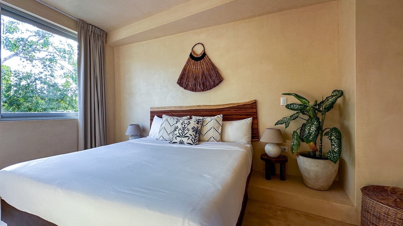 Bedroom with a king bed, carved wood headboard, patterned pillows, and a window overlooking trees.