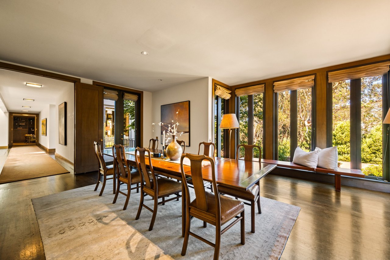 Dining room with a long wooden table, upholstered chairs, and expansive garden-facing windows.