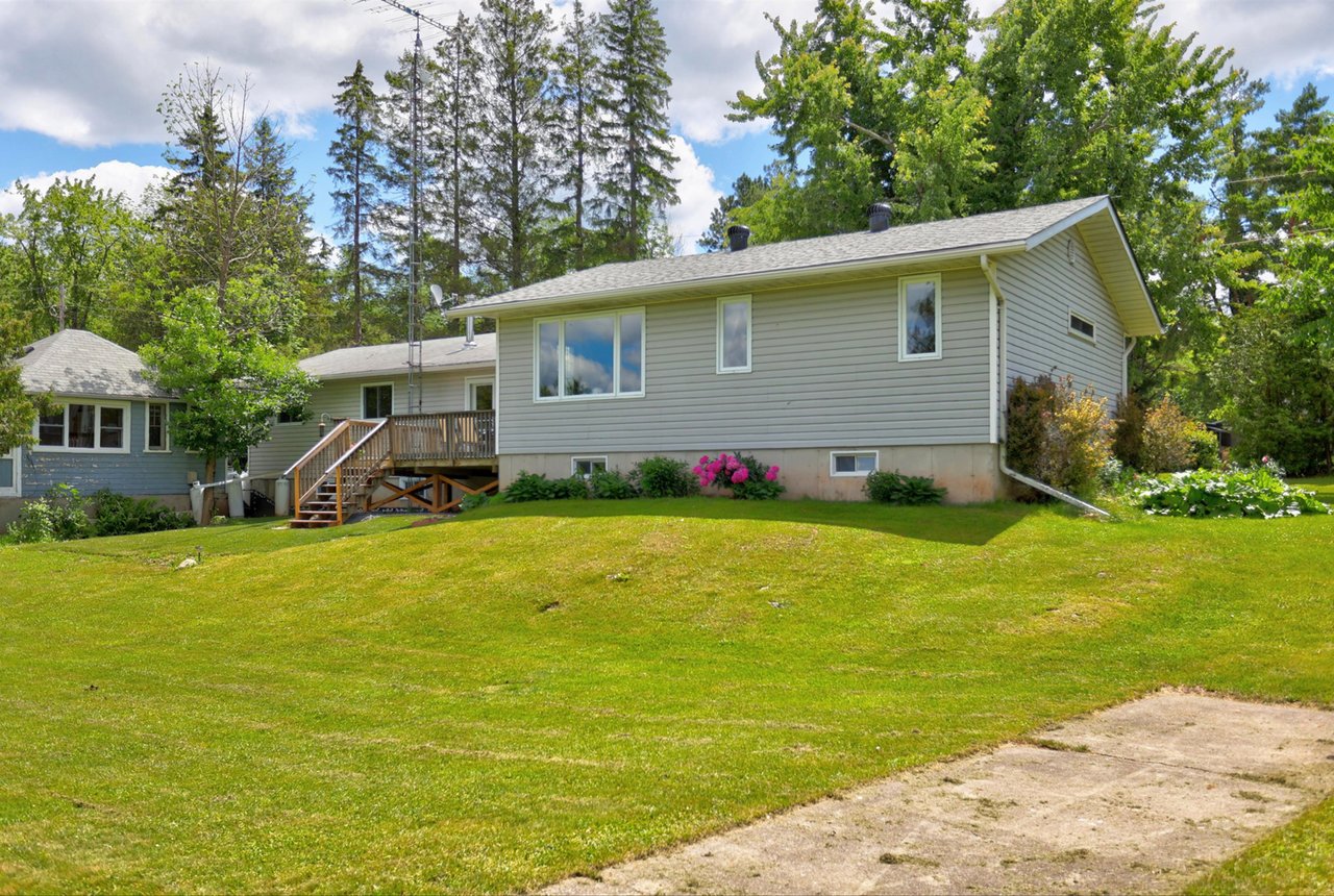 Gray-sided house with a raised wooden deck and stairs beside a broad lawn and mature trees.