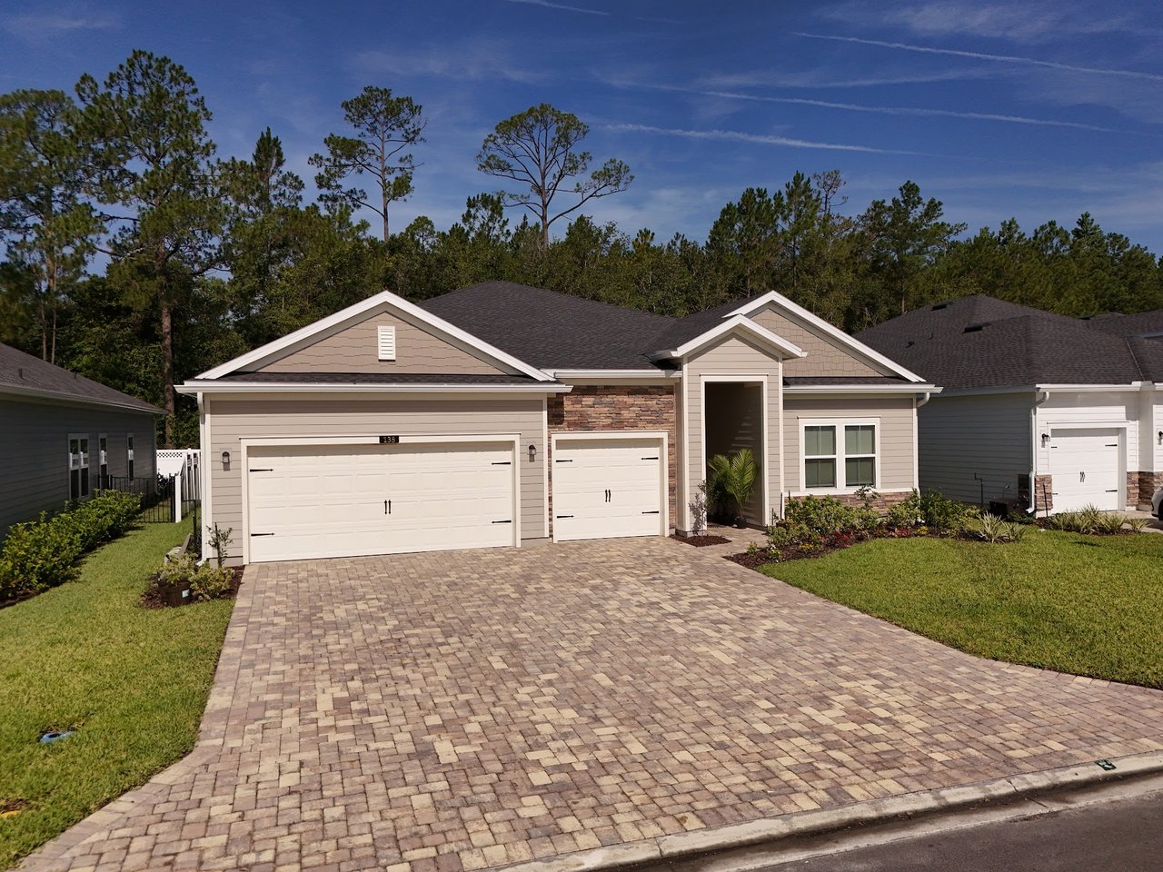 Beige single-story home with two garage doors, stone accents, and a wide paver driveway.