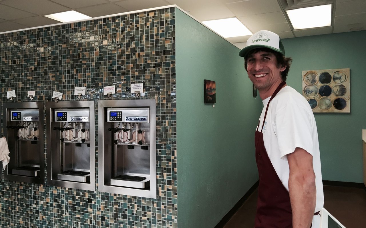 Frozen yogurt shop employee smiling beside three frozen yogurt machines.