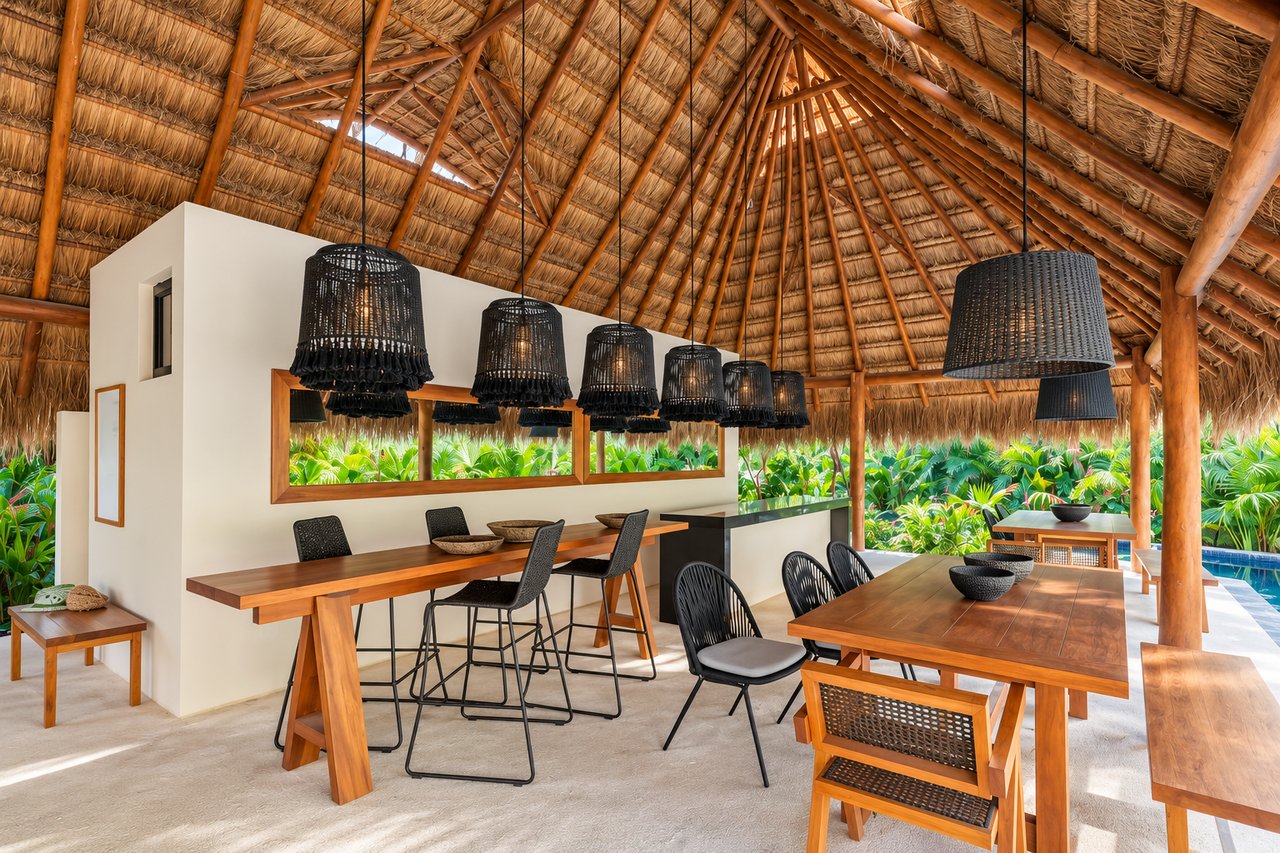 Open-air dining area beneath a tall thatched roof, with wooden tables and tropical greenery beyond.