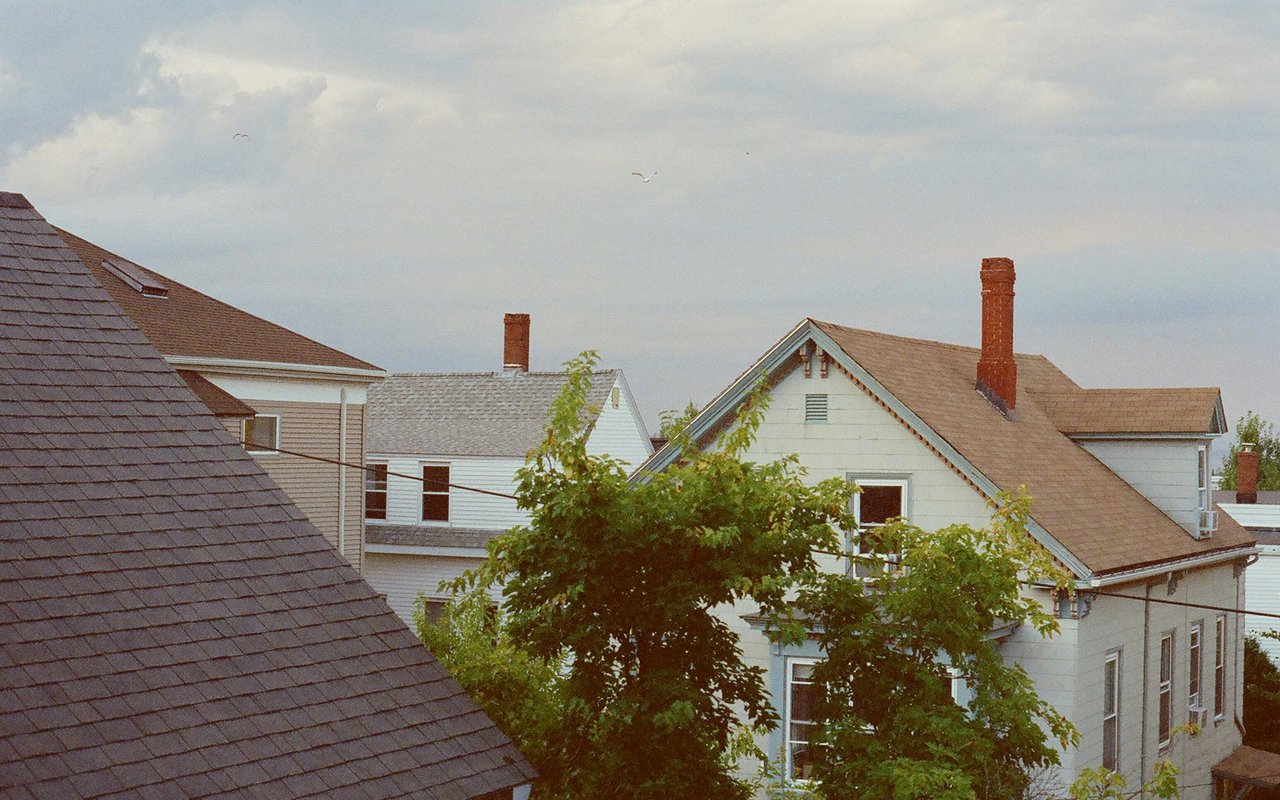 Town rooftops with brick chimneys and leafy trees beneath a cloudy sky.