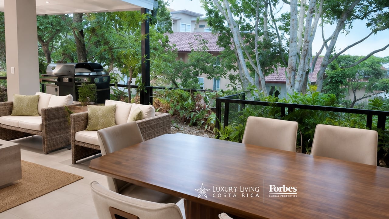 Covered terrace with a wooden dining table, wicker lounge seating, and a barbecue overlooking leafy greenery.