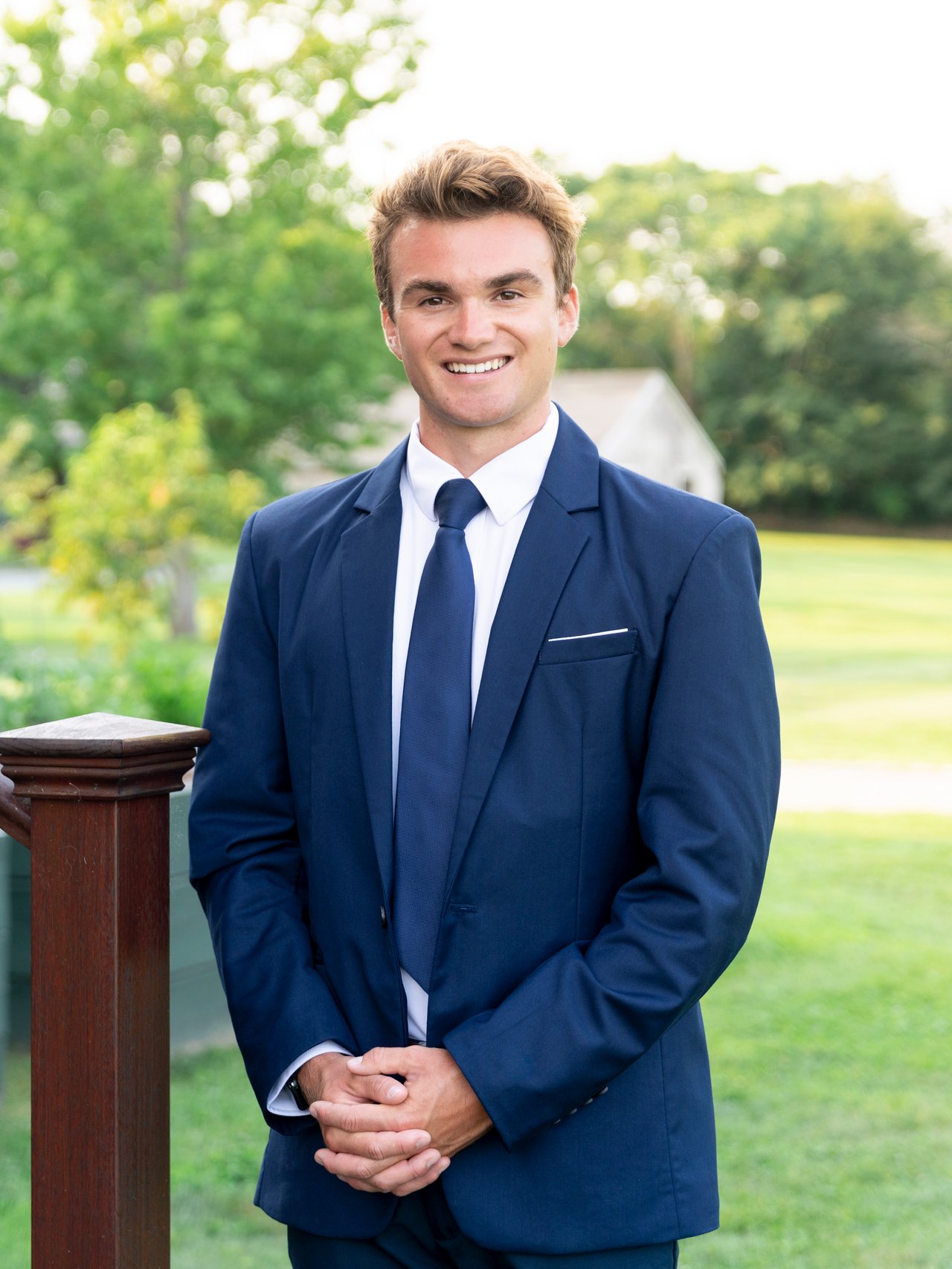 Nick Peters in a navy suit and tie, posing outdoors against green trees and lawn.