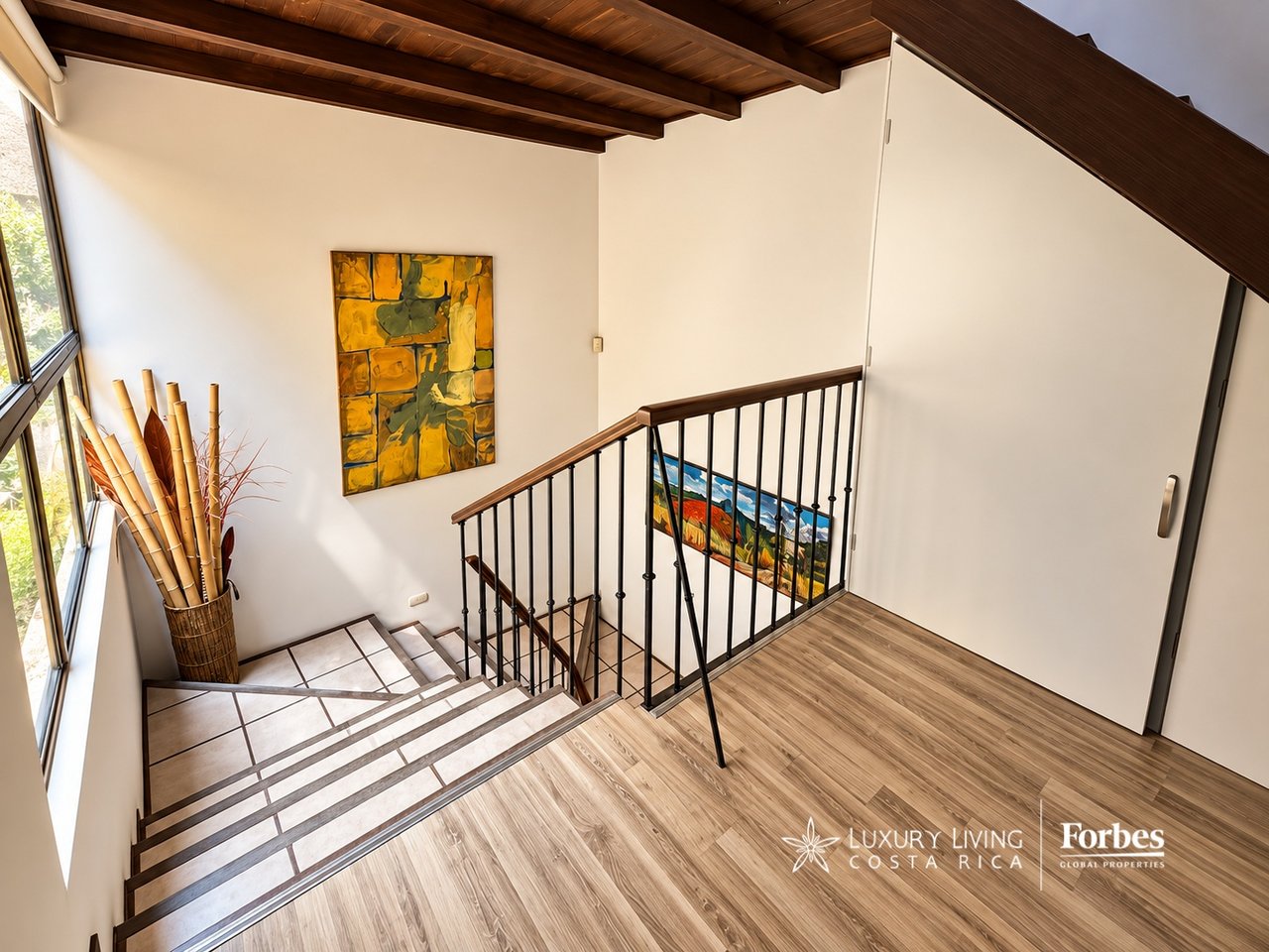 Home stairwell with tiled steps, dark railings, wood ceiling, large windows, and colorful artwork.