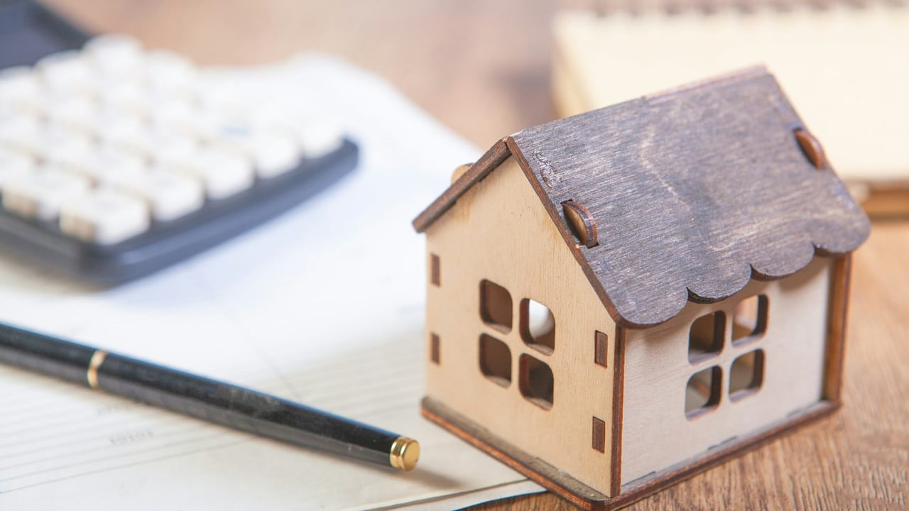 Small wooden model house beside a calculator and pen resting on paperwork on a wooden desk
