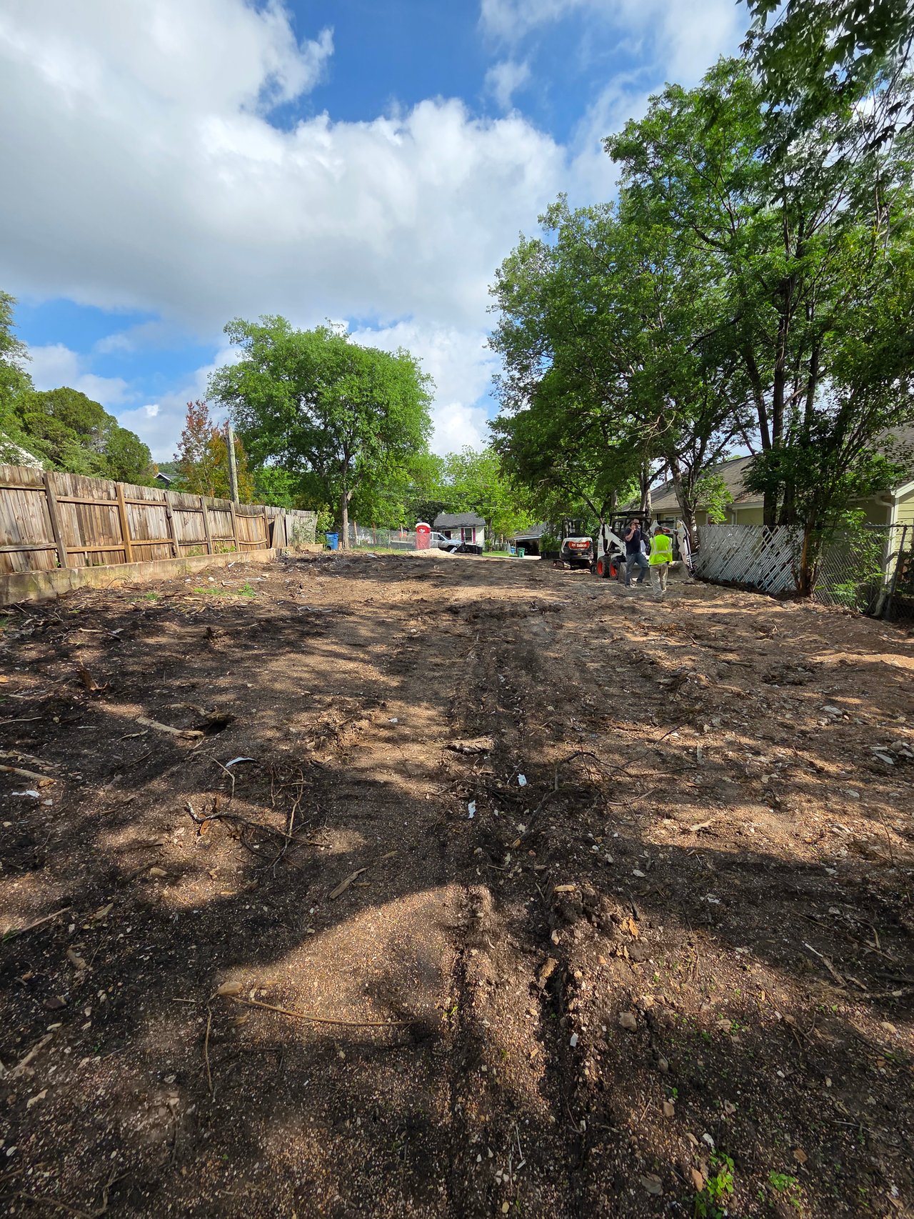 Cleared development lot with workers and construction equipment between fences and mature trees.