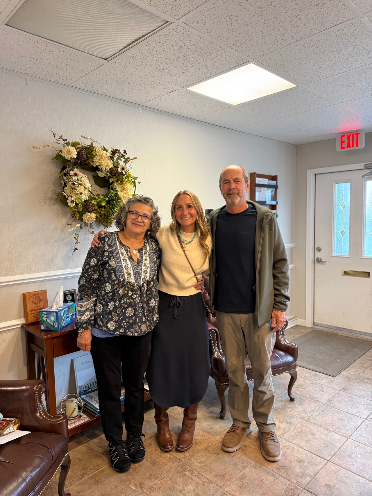 Three adults pose together in an office reception room beneath a floral wreath.
