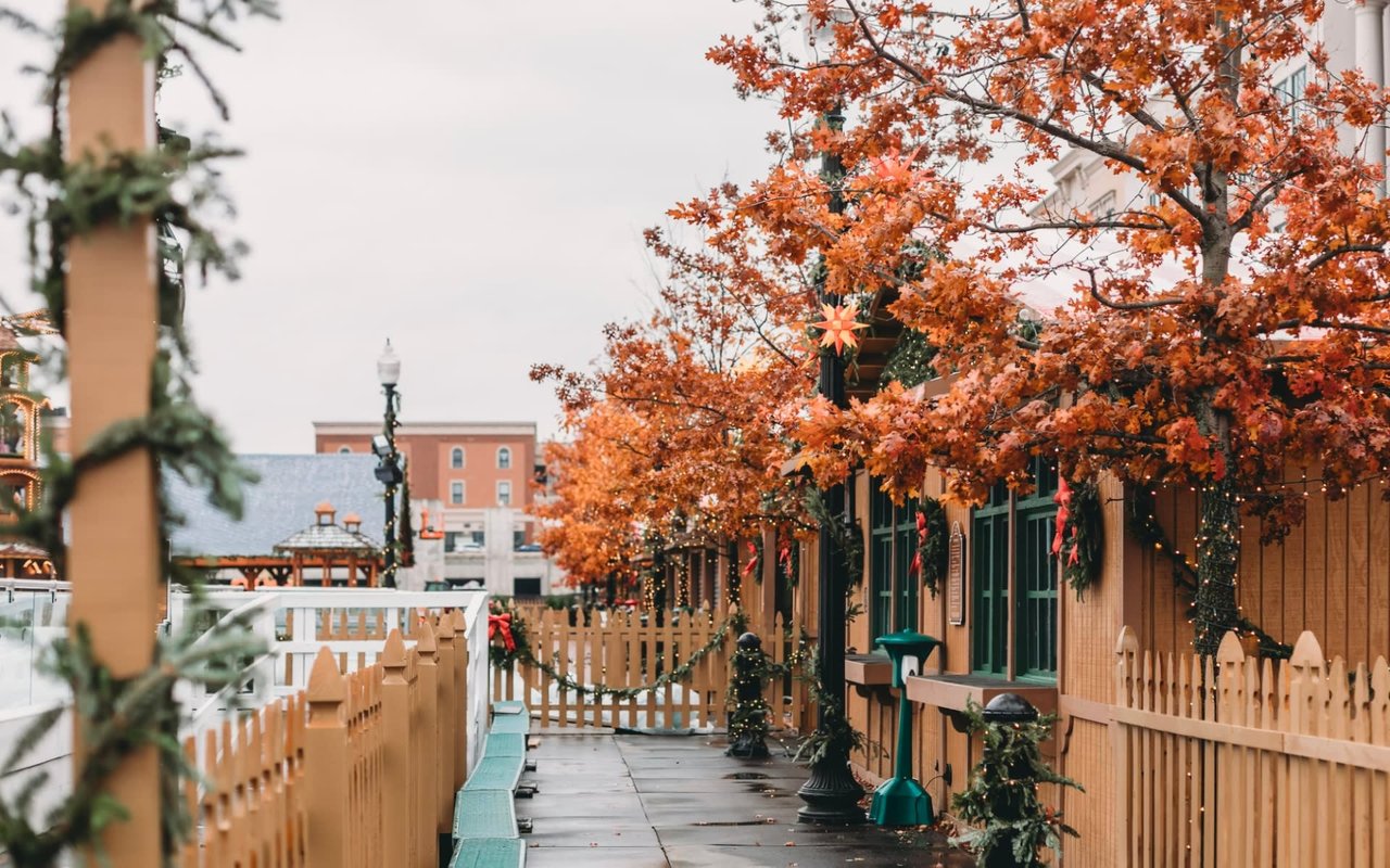 Vibrant autumn view of the Carmel Christkindlmarkt featuring wooden German-style vendor huts, festive wreaths, picket fencing, and seasonal fall foliage.