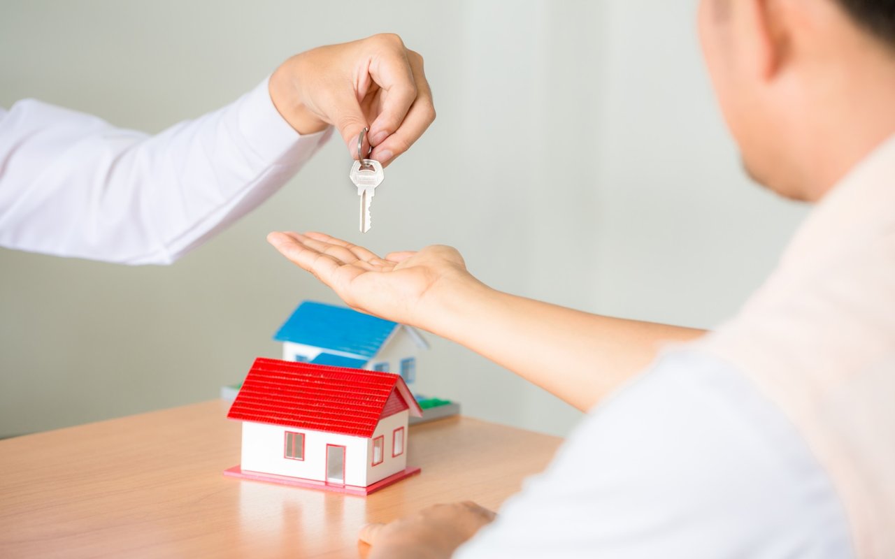 A person hands a house key to another beside two model homes.