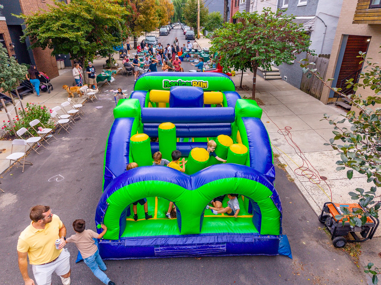 Neighbors gather around a large blue-and-green inflatable obstacle course at a residential street party.