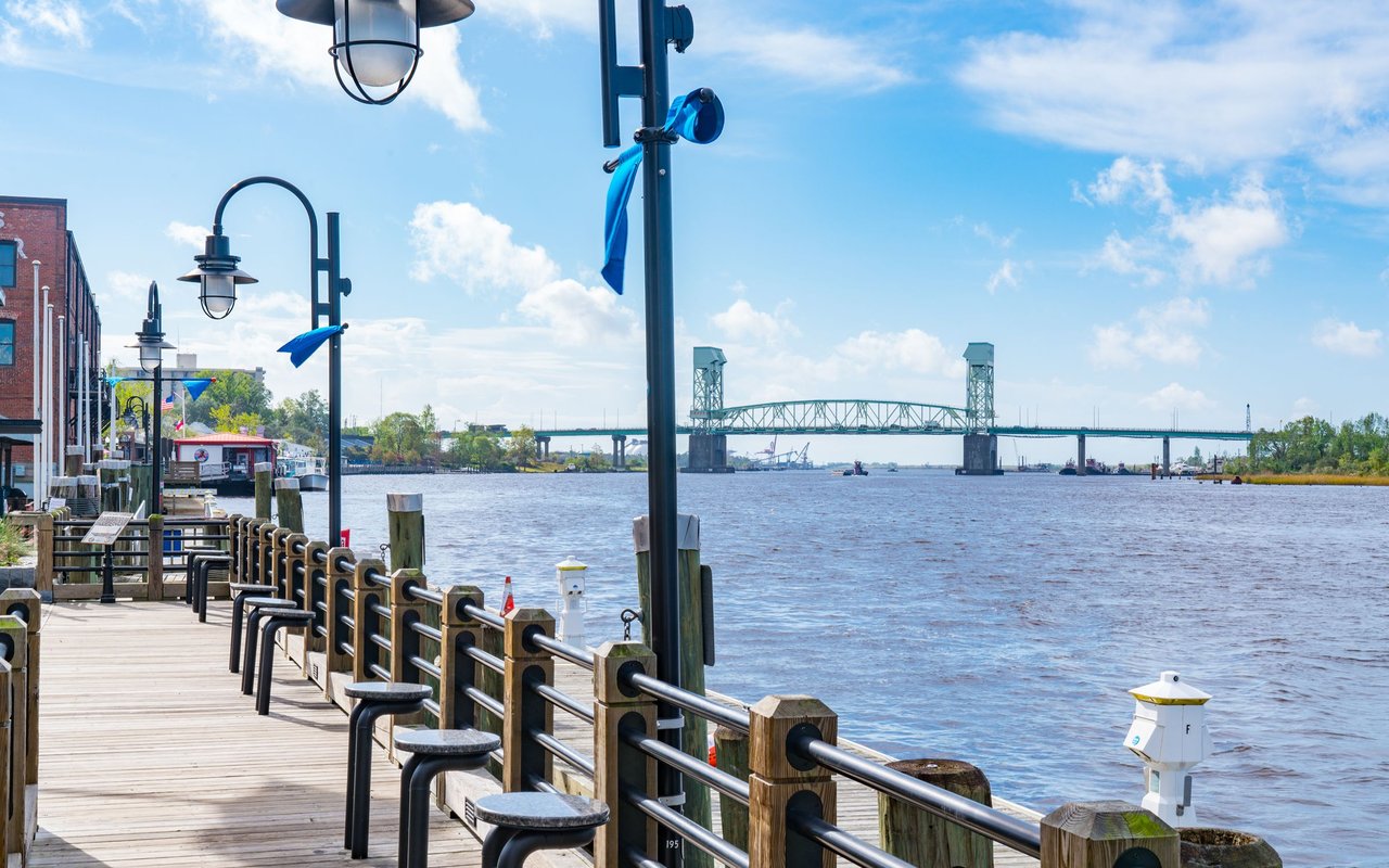 Wilmington riverfront boardwalk beside the Cape Fear River, with a steel bridge in the distance.
