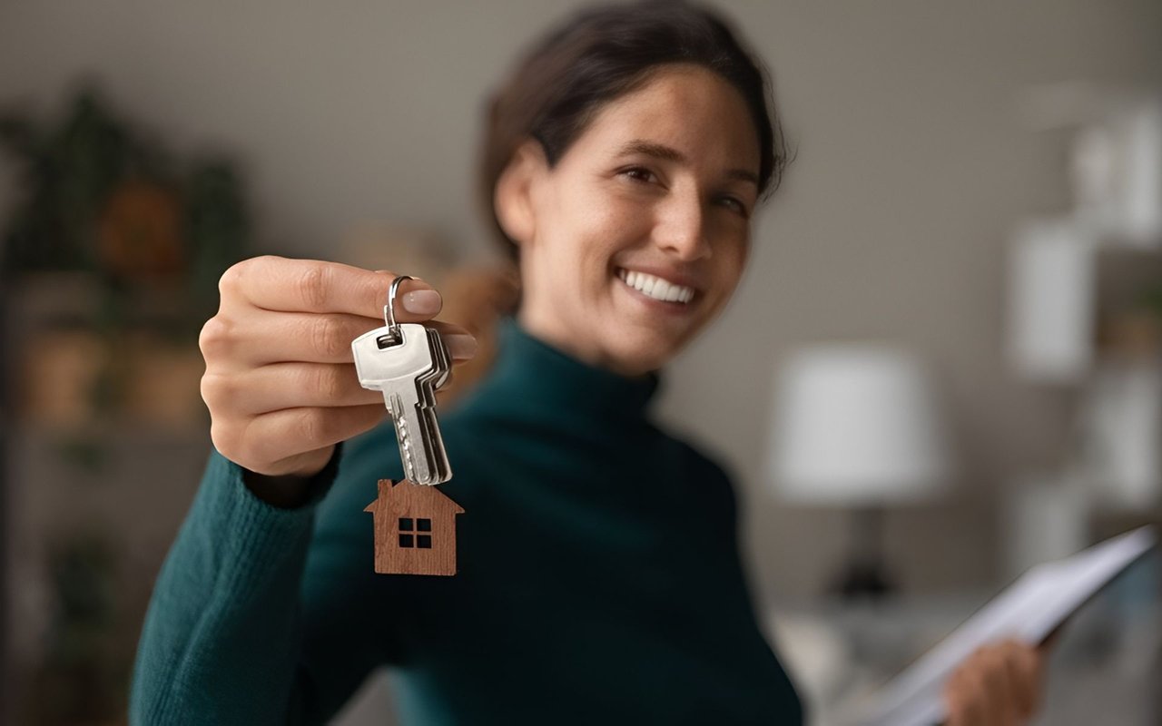 Smiling woman holds up house keys with a wooden house-shaped keychain.