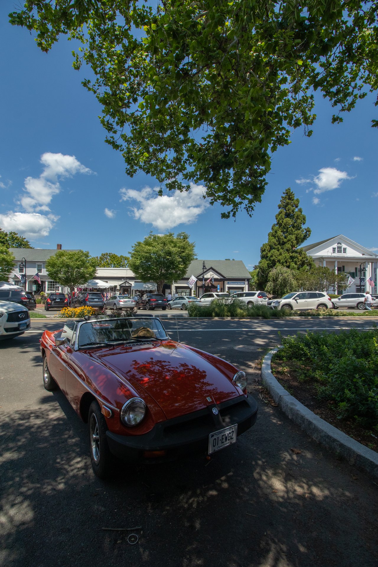 Red vintage MG convertible parked along a sunny village street with shops and leafy trees.
