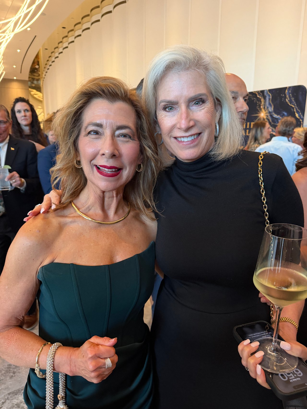 Two women pose together at a reception, one in a teal dress and the other in black.